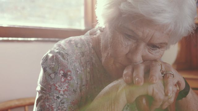 Senior woman thoughtfully leaning on walking cane, bathed in natural light from window. Ideal for topics on aging, contemplation, senior care, domestic life, and health awareness. Suitable for articles, caregiving services, and wellness content.