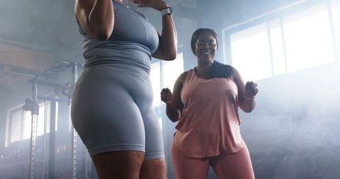 Plus-size women working out together in industrial gym, backlit window haze, activewear