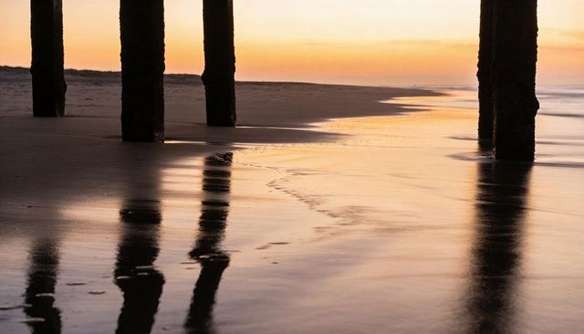 Sunset light casting long reflections beneath weathered pier pilings on wet sand