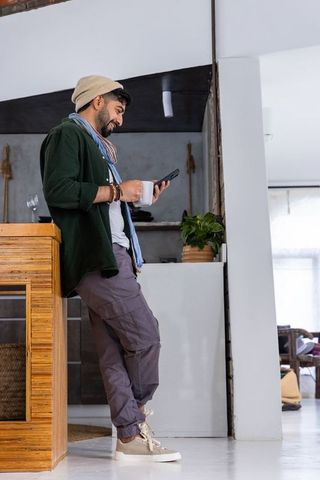 Casual man leaning on kitchen island holding smartphone and coffee mug in modern home