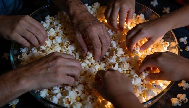 Multigenerational hands reaching for popcorn in large glass bowl during cozy movie night