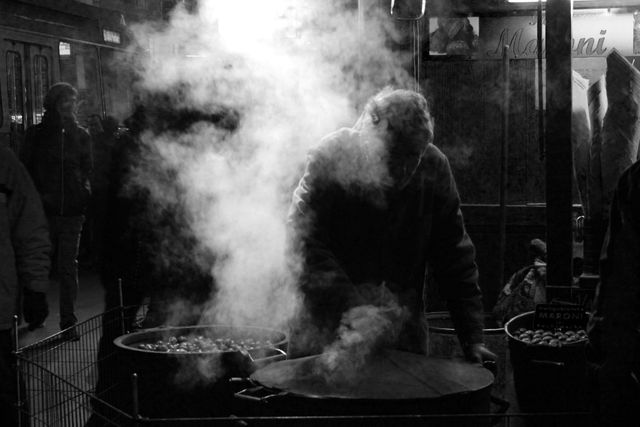 Street vendor roasting chestnuts at night, shrouded in smoke, creates atmospheric and traditional scene. Ideal for use in articles about street food culture, urban night markets, or traditional winter snacks. Can also be used for describing city life and street vendors.