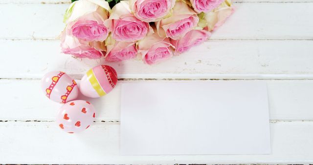 A collection of pastel-colored roses and decorated Easter eggs are arranged next to a blank card on a white wooden surface, with copy space. It suggests a festive Easter celebration or a spring-themed greeting card opportunity.