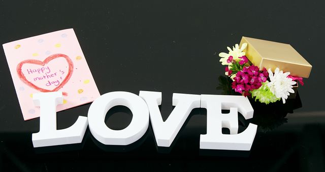 A Mother's Day card, a bouquet of flowers, and the word LOVE in white letters are arranged on a black surface, with copy space. These items symbolize affection and celebration for Mother's Day.