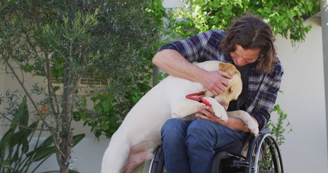 A man in a wheelchair is embracing a Labrador dog while sitting outdoors surrounded by greenery. The man appears to be very affectionate and connected with his dog, which adds to the warm, heartfelt moment. This image is suitable for use in topics related to companion animals, disability advocacy, therapy animal programs, and the emotional bond between pets and their owners.