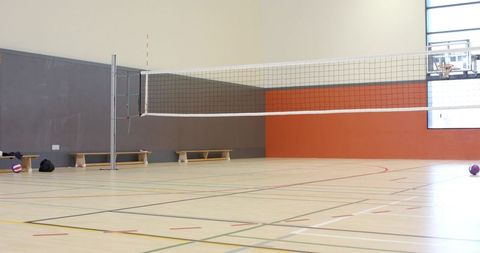 Empty sunlit indoor volleyball court with net, benches and basketball hoop in gymnasium