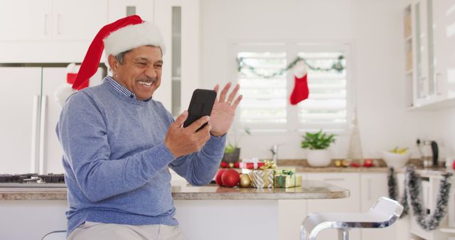 Senior man wearing Santa hat, sitting in kitchen, using smartphone for video calling during Christmas holidays. Ideal for content about festive family connections, holiday greetings, and elderly embracing technology. Can be used for advertisements, blogs, social media posts, and promotional material related to Christmas celebrations, seasonal greetings, and family interactions.