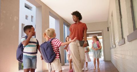 African american teacher walking hallway guiding primary students with backpacks