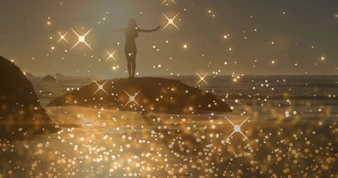 Woman standing on coastal rock at sunset with golden bokeh and starburst flares