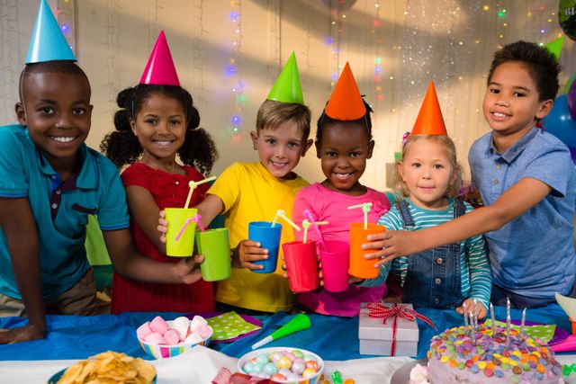 Children of diverse backgrounds are celebrating a birthday party, toasting with colorful drinks while wearing party hats. They are standing around a table with a cake, gifts, and various party decorations. This image can be used for themes related to childhood, celebrations, diversity, friendship, and festive occasions.