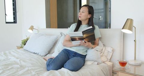 Chinese woman reading paperback on bed in minimalist bedroom, cozy daylight lifestyle