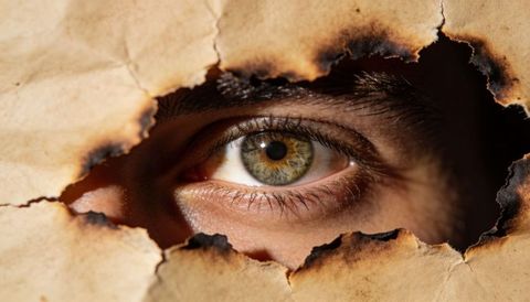 Peering green-hazel eye through burned paper hole with charred edges dramatic close-up