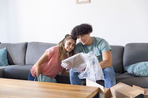 Young couple unpacking new home on sofa inspecting bubble-wrapped item and moving boxes