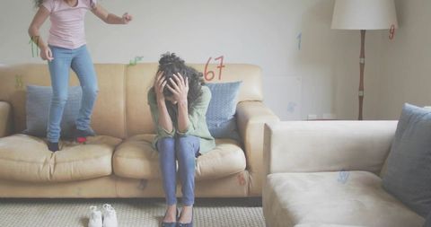 Overwhelmed mother covering face while child jumping on couch in cluttered living room