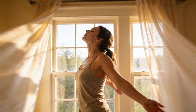 Sunlit woman inhaling deeply and stretching at window with flowing sheer curtains