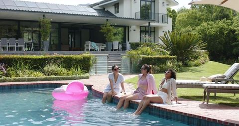 Three friends relaxing poolside at modern luxury villa with pink float and summer swimwear