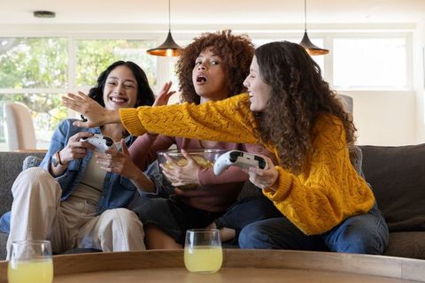 Multicultural Friends Laughing and Gaming on Couch Reaching for Controller and Snacks