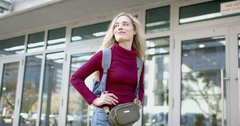 Young woman standing with backpack and olive crossbody bag looking up at glass entrance