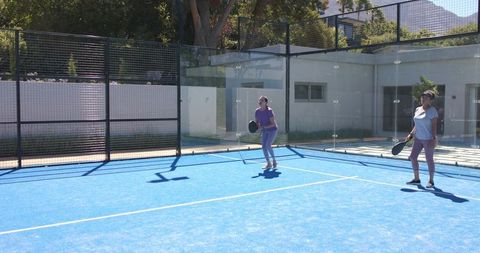 Women playing padel on blue court with glass walls, sunny outdoor recreational sport