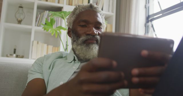 Elderly man with grey beard relaxing on couch while reading or browsing on digital tablet at home. Ideal for depicting leisure time, use of modern technology by seniors, and relaxed home environment settings.