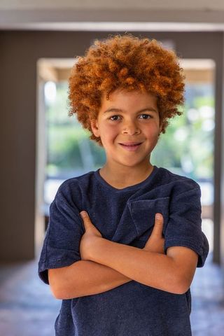 Confident red curly-haired boy posing arms crossed in sunlit home doorway