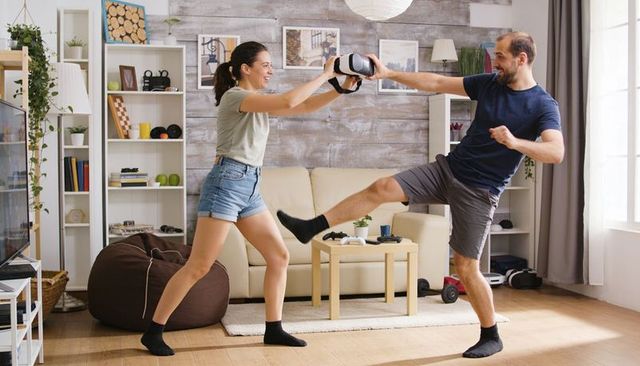 Couple sharing vr headset and playfully kicking during bright cozy living room gaming