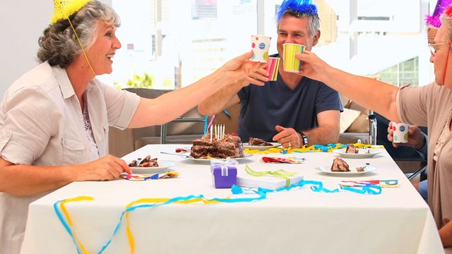 Elderly friends gathering at home for a birthday celebration, wearing party hats and raising cups in a toast. A decorated table with cake and party supplies suggests a light-hearted, joyful occasion. Suitable for promoting social gatherings, celebrating friendship, lifestyles of the elderly, positive aging, and party supplies.