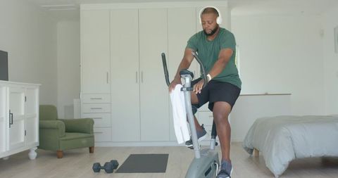 African man exercising on stationary bike in modern bedroom with headphones and dumbbells