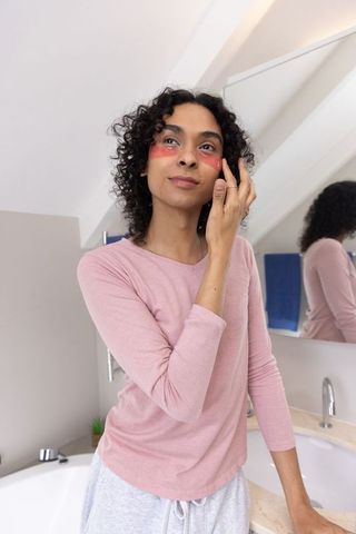 Non-binary person adjusting under-eye gel patches at bathroom vanity during calm self-care