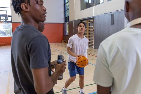Diverse male teammates practicing basketball in sunlit indoor gym holding ball