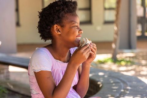 African american girl laughing and eating sandwich on school courtyard bench in sunlight
