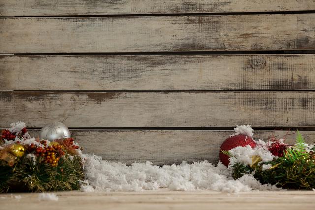 Rustic wooden background decorated with fake snow and Christmas ornaments, including wreaths with berries and pine, perfect for holiday greeting card designs, festive marketing materials, or winter-themed displays.