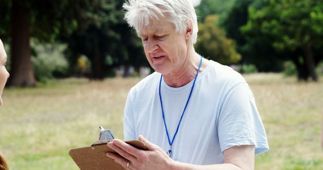A senior Caucasian man is engaged in a conversation with an individual, holding a clipboard and pen, in a role such as a volunteer or surveyor. His focused expression suggests he is attentively listening or recording information during an outdoor event or activity.