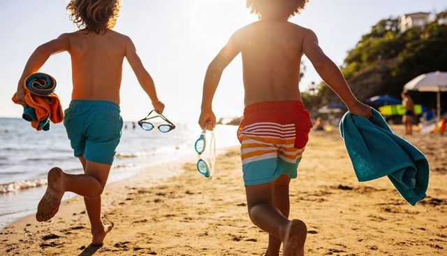 Running young boys on sunlit beach carrying colorful towels and goggles during golden hour