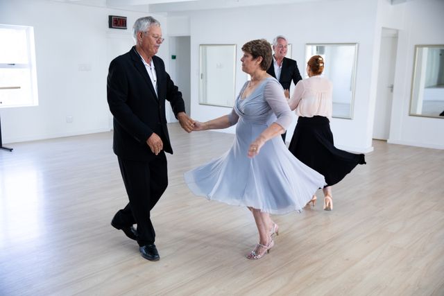 Two happy elegant Caucasian senior couples spending time together in a ballroom, taking part in dancing class, dancing, holding hands and smiling. Active golden years hobby.