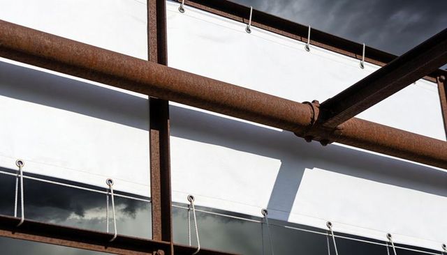 Rusted steel beams crossing white panels, grommets and ropes revealing glass strip