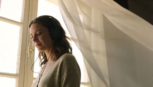 Woman listening with wired headphones by sunlit multi-pane window and billowing curtain