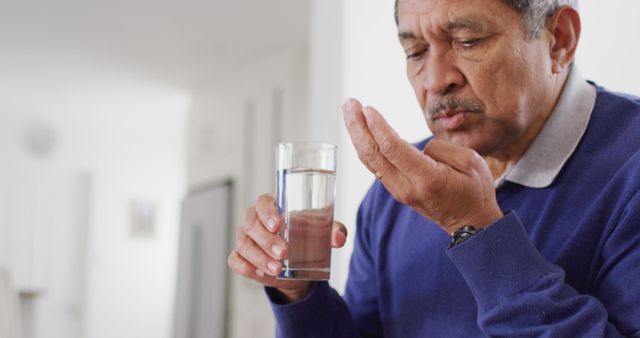Image of senior biracial man taking pills and drinking water at home. Retirement, medicine, healthcare, inclusivity and senior lifestyle concept.