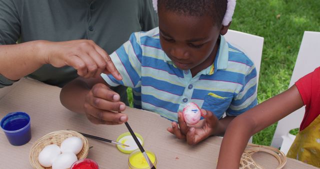 Happy african american boy painting easter eggs with family in garden. family easter, togetherness, celebration and spending quality time concept.