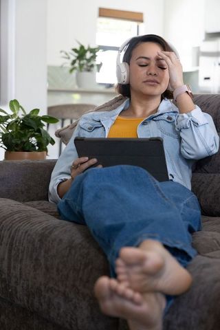 African american woman relaxing on couch wearing headphones holding tablet feeling mindful