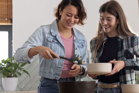 Friends cooking in cozy home kitchen serving pasta into ceramic bowl smiling together