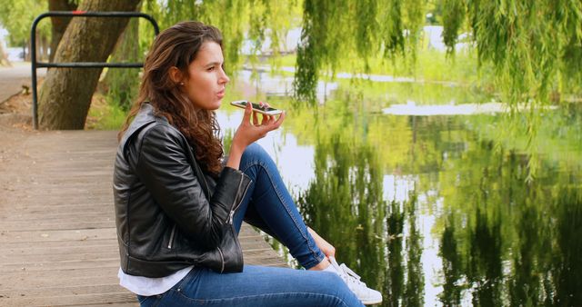 A young Caucasian woman enjoys a snack while sitting by a serene lake, with copy space. Her casual pose and contemplative gaze suggest a moment of relaxation amidst natural surroundings.