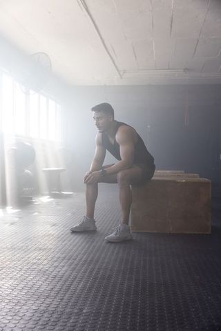 Hispanic athlete resting on plyometric box in sunlit gym after intense strength workout