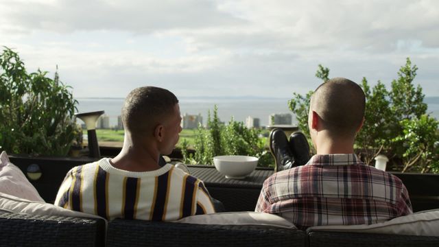 Two men sit and enjoy their time together on a roof terrace with an expansive view of the skyline under a cloudy sky. This moment of camaraderie and relaxation can be used for social connection or lifestyle imagery in promotional materials, articles about urban living or scenes promoting casual leisure activities.