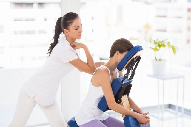 Masseuse providing a back massage to a woman seated on a massage chair in a bright, serene spa environment. Ideal for use in wellness and health-related content, spa advertisements, therapy services promotions, and articles about stress relief and relaxation techniques.