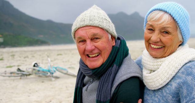 Elderly man and woman are smiling while experiencing a joyful moment at the beach during winter. They are dressed warmly in knitted hats and scarves. This is perfect for content related to senior lifestyles, winter outdoor activities, retirement, and the positive aspects of aging. It can also promote health and wellness for older adults.