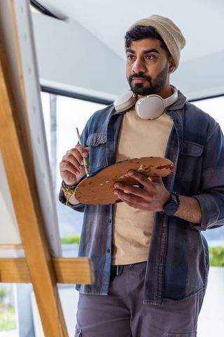 Artist painting at easel holding palette and brush wearing beanie and headphones in bright studio
