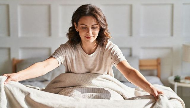 Middle-aged woman making bed in sunlit bedroom smoothing cozy duvet for peaceful morning routine