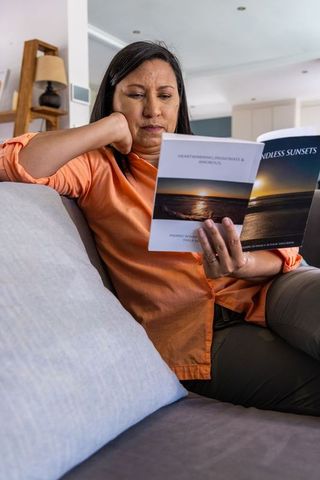 Mature woman reading sunset-cover paperback on gray sofa wearing orange shirt for leisure