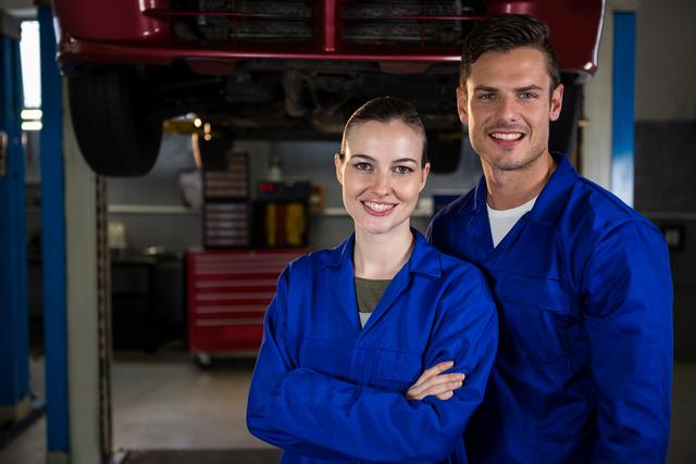 Two mechanics in blue uniforms standing under a car in a repair garage, smiling confidently. Ideal for use in automotive service advertisements, repair shop promotions, and articles on car maintenance and professional teamwork.